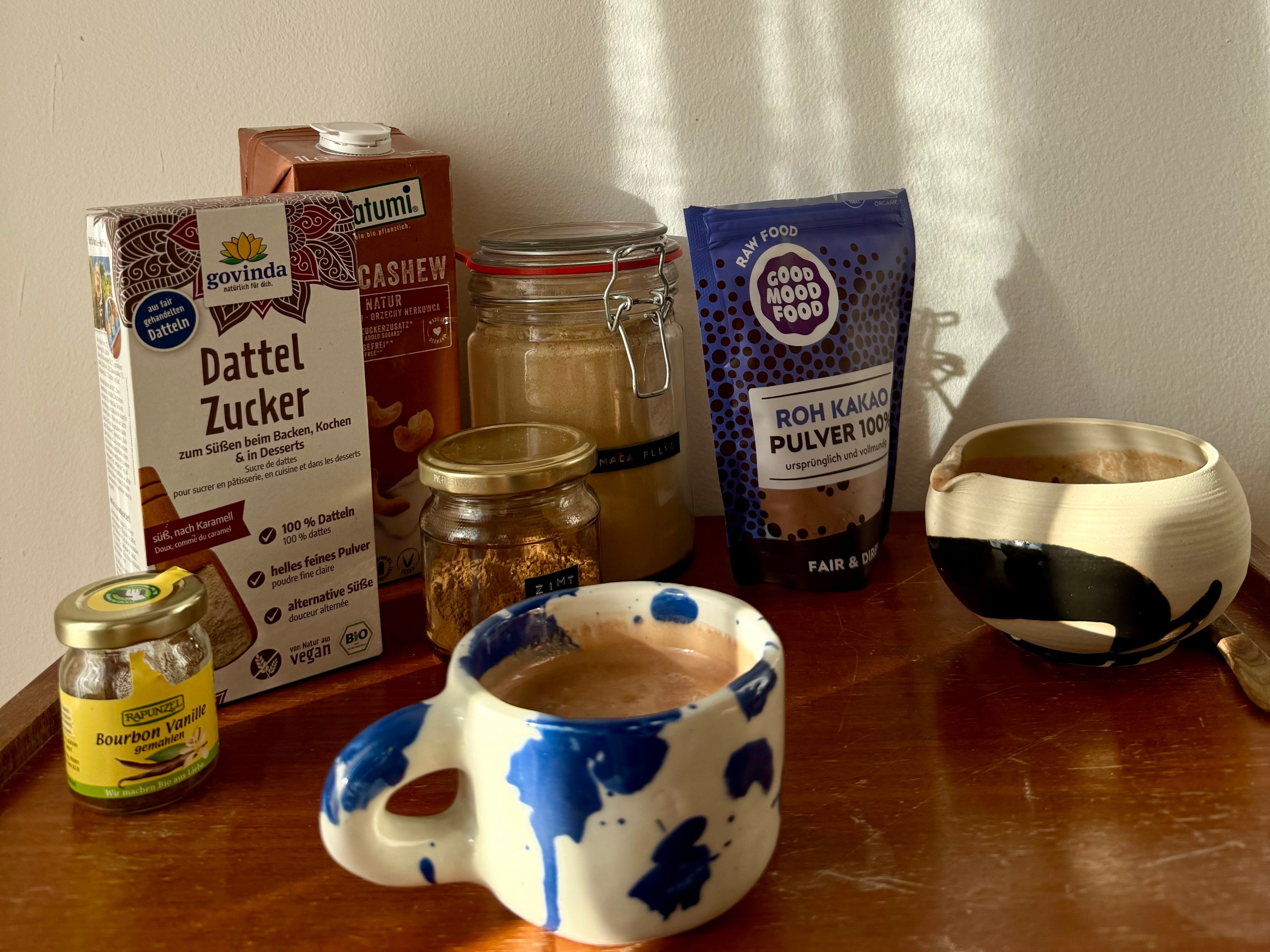 Warmer Raw Superfood Cacao in einer hellen Keramik-Tasse, umgeben von Rohkakao, Zimt und Maca – nährstoffreicher Wohlfühl-Drink für Schwangerschaft und Stillzeit